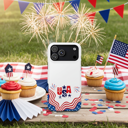 A white smartphone case with a patriotic red, white, and blue design stands on a wooden table outdoors. The scene includes cupcakes, small American flags, confetti, bunting, and fireworks in the background, making this a lifestyle shot.