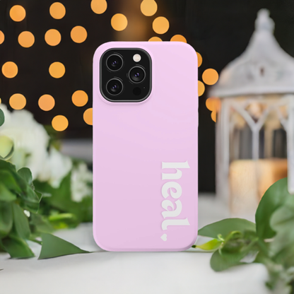 A pink iPhone case with the word 'heal' written on it, displayed against a backdrop of white lanterns and greenery.